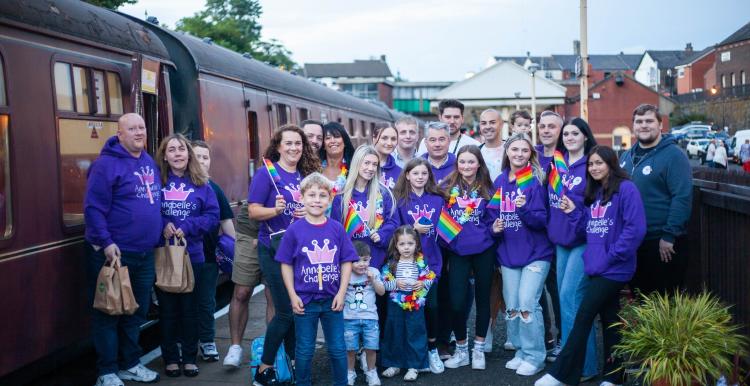 A group of people wearing purple hoodies standing on a railway platform holding rainbow flags.