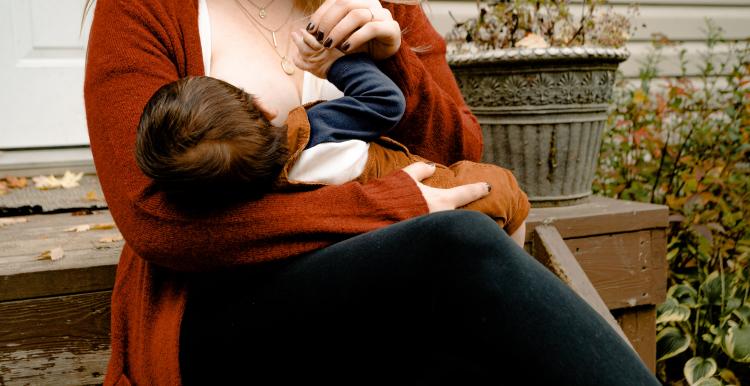 A woman sitting on a bench breastfeeding her baby. The baby as dark brown hair and the woman is wearing an orange cardigan.