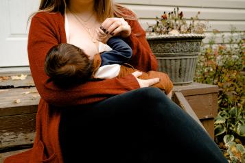 A woman sitting on a bench breastfeeding her baby. The baby as dark brown hair and the woman is wearing an orange cardigan.